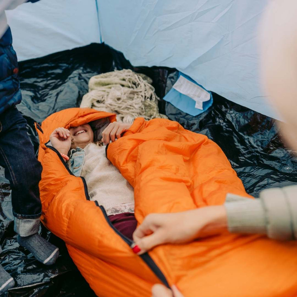 A child sleeping in an orange camping mummy bag inside a tent, with an adult's hand opening the zipper. Another person's legs are visible at the side of the tent.