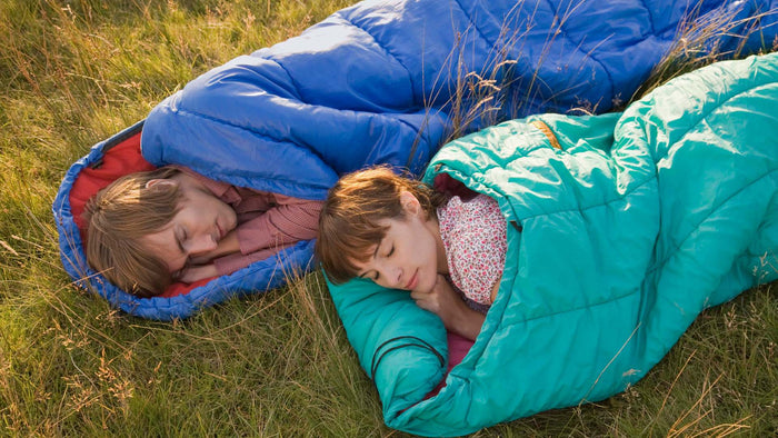 Two children sleeping in colorful camping sleeping bags on a grassy field at sunset. One sleeping bag is bright blue with a red lining, and the other is teal with a pink patterned lining.