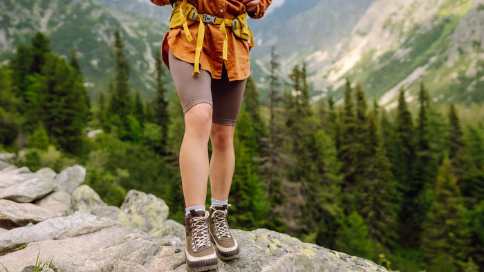 A person hiking on a rocky mountain path wearing brown biker shorts, an orange-brown jacket, hiking boots, and a yellow harness, with a lush green forest and mountains in the background.