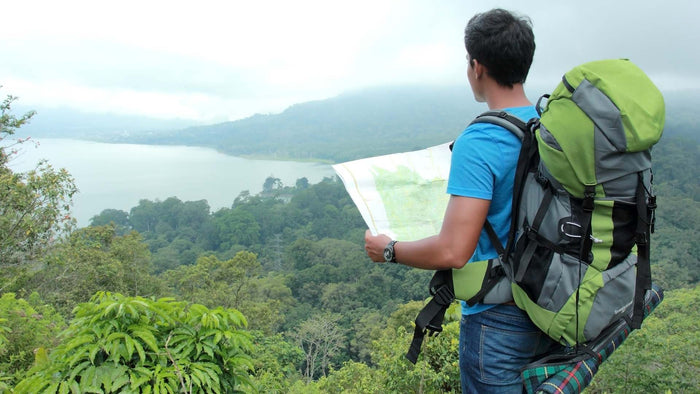 A man wearing a large green and gray hiking backpack stands on a forested mountaintop, holding a paper map with a lake and misty mountains in the background.