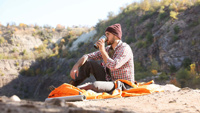 A man sitting on a rocky cliffside campsite wearing a red and white plaid shirt, dark pants, and a maroon beanie, drinking from a metal can. An orange tent is laid out on the ground in front of him, with a rocky, forested mountain slope in the background.
