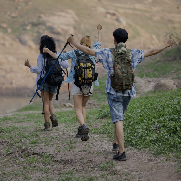 Three young hikers with backpacks and trekking poles walk arm-in-arm along a dirt path with a scenic river and rocky hillside in the background.