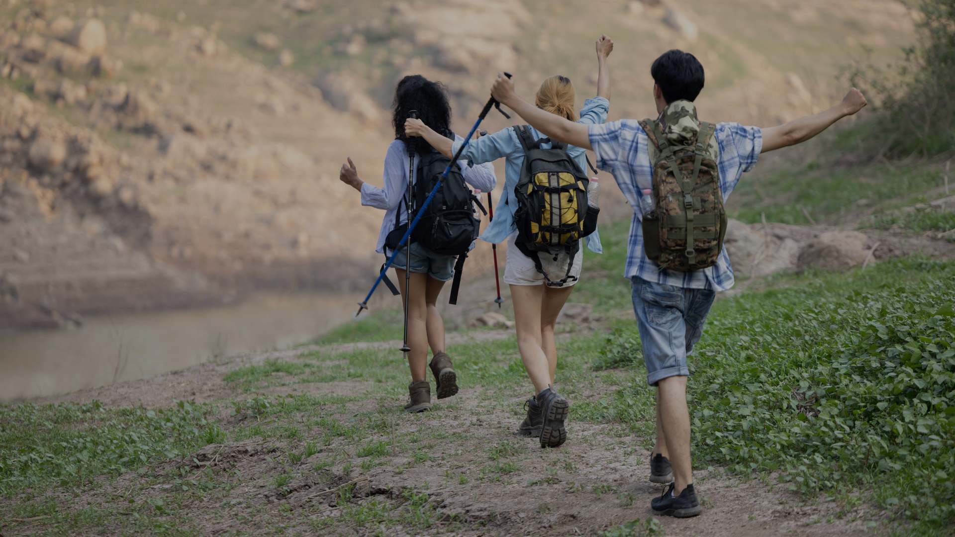 Three young hikers with backpacks and trekking poles walk arm-in-arm along a dirt path with a scenic river and rocky hillside in the background.