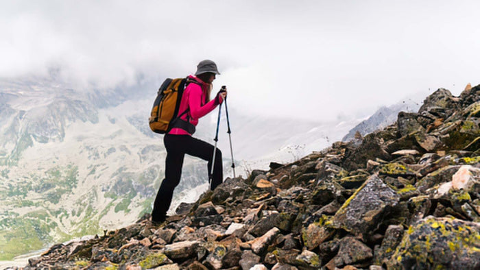 A hiker wearing a bright pink jacket, black pants, and a gray helmet carries a large brown and orange backpack while using trekking poles to ascend a rocky mountain ridge with misty peaks in the background.