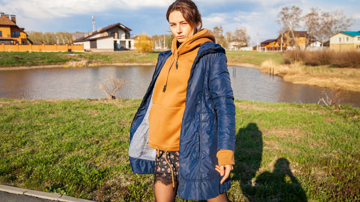 A woman standing outdoors near a grassy field and a small pond, wearing a long navy blue puffer coat over a mustard yellow hoodie, with patterned shorts visible underneath.